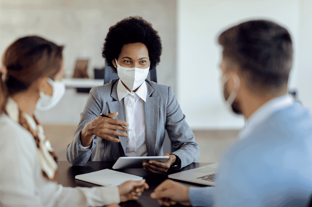 Matéria MBD - Cheque Especial - Consultor financeiro afro-americana conversando com um casal, usando máscara facial protetora.